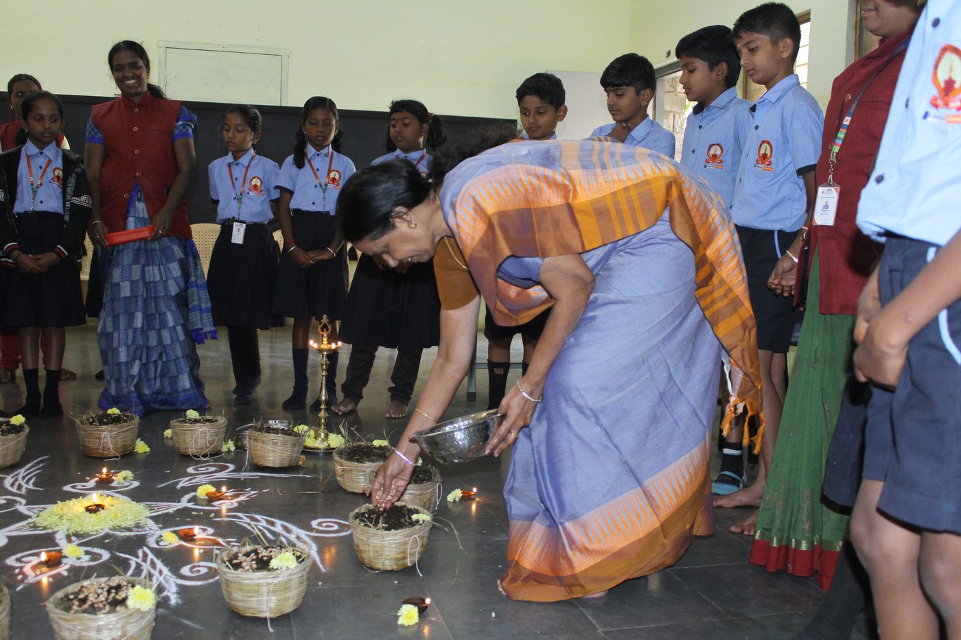 Mrs. Prema Meru (Amma) sowing seeds in baskets at a lamp-lit ceremony while students and teachers look on