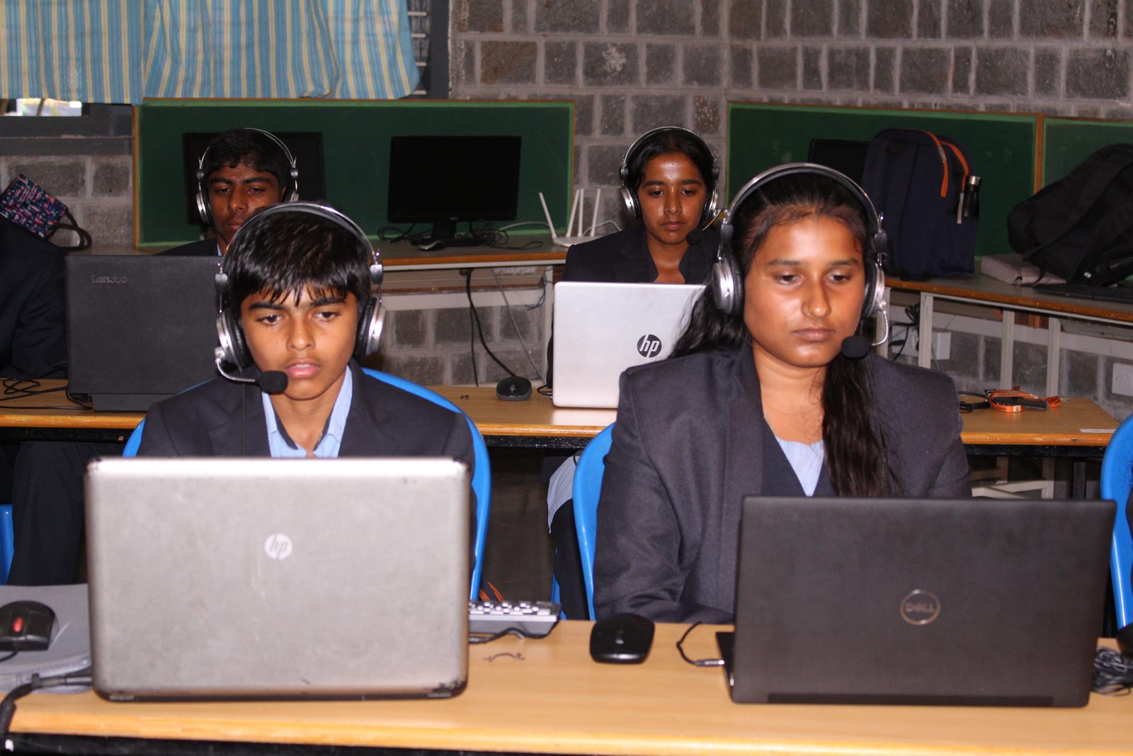 Two students with headsets at their laptops, with more students behind