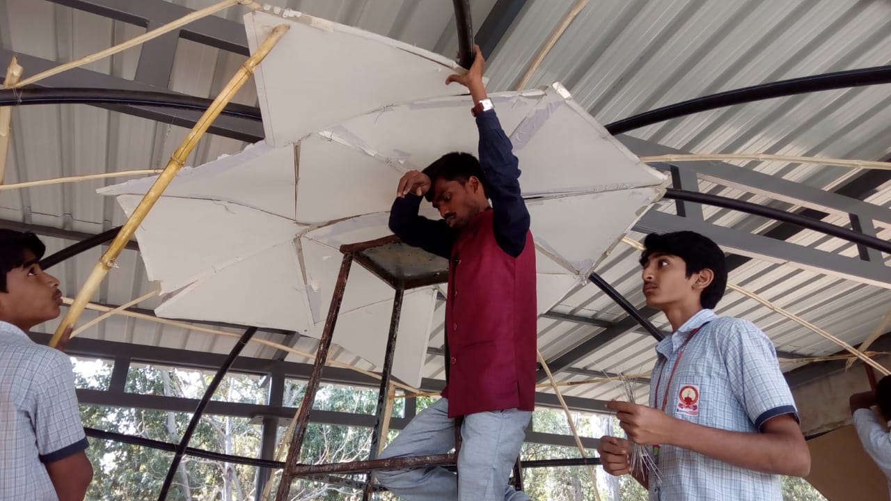 A teacher fitting angular cardboard panels near the apex of the dome while students look on