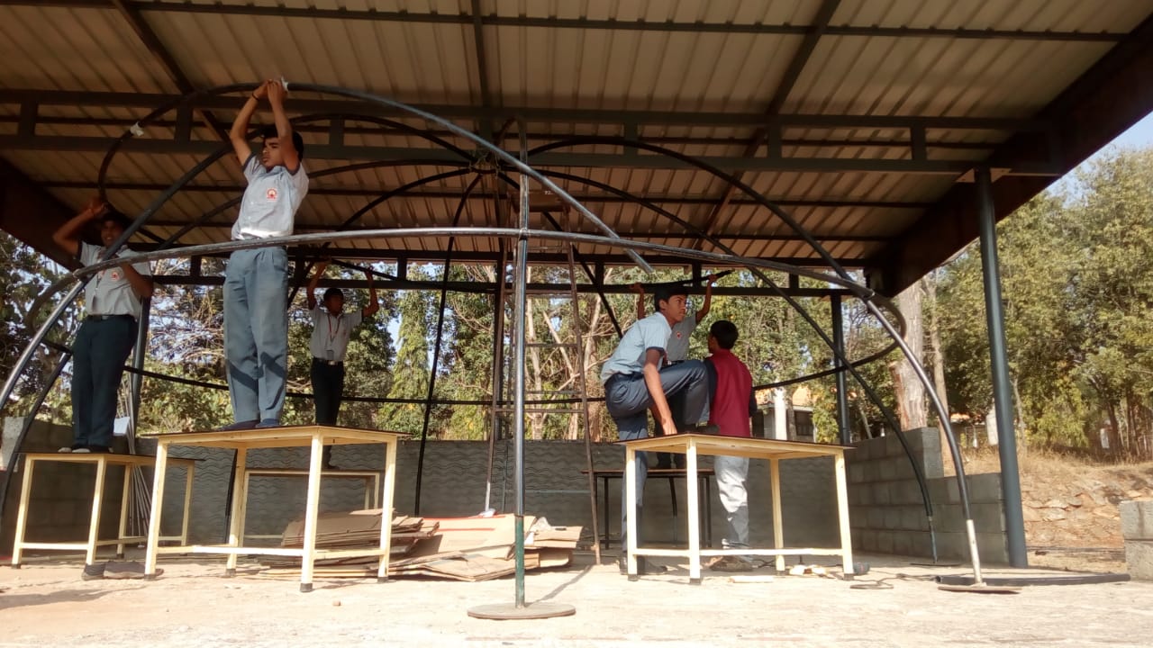 The nearly completed geodesic frame of the Version 2 planetarium dome, with students balanced on desks tying off the final ribs