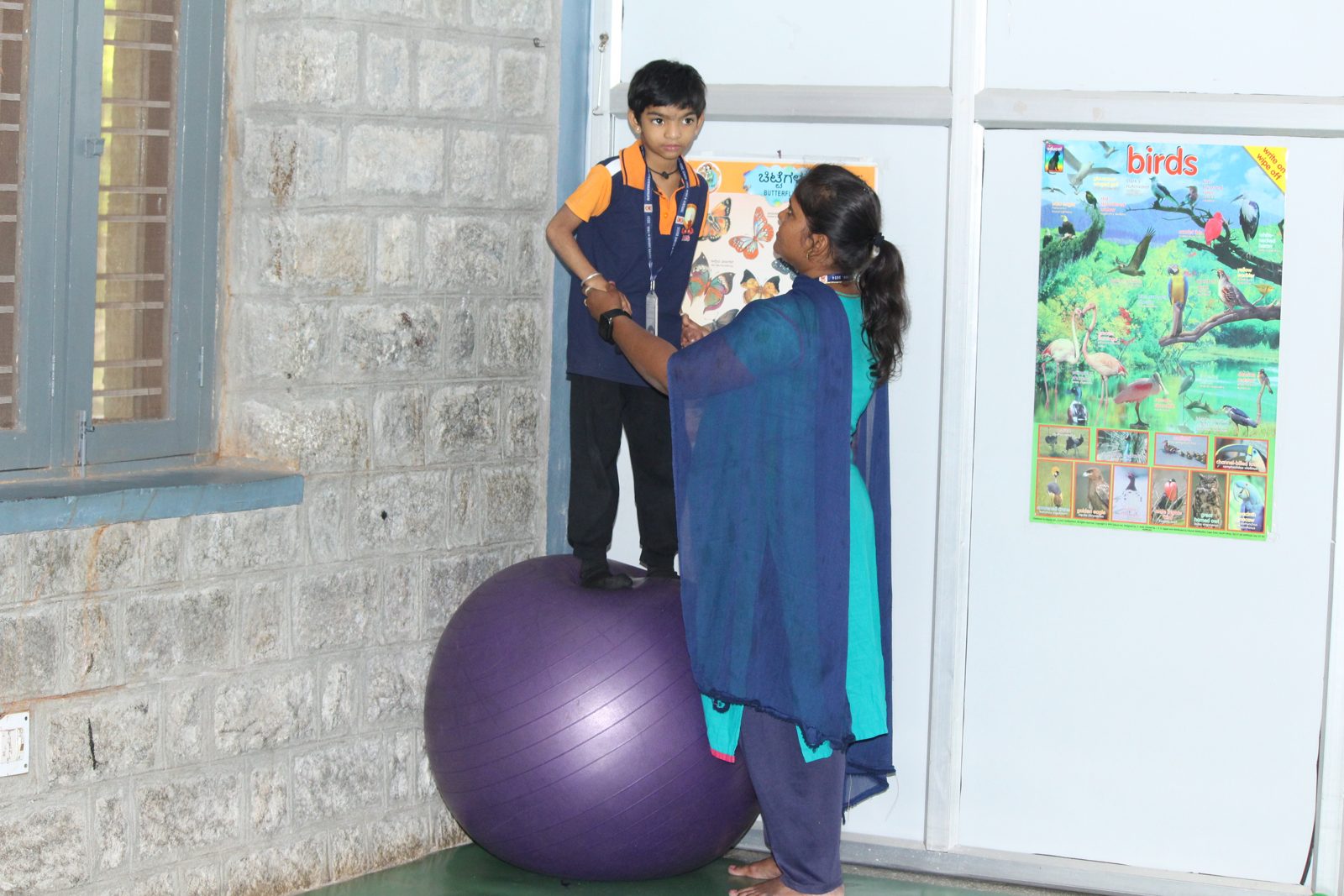A child standing on a large therapy ball with an educator holding on for support