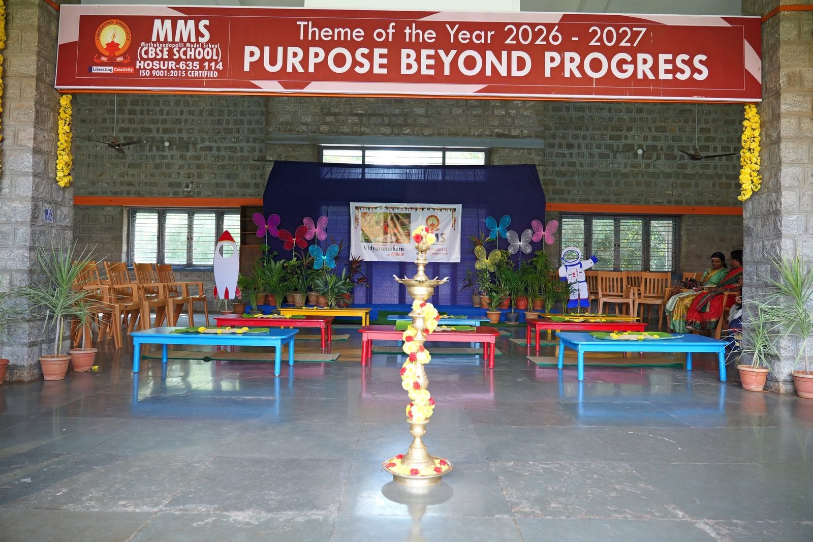 The decorated Vidhyarambam hall at MMS with a lit lamp and the Theme of the Year 2026-2027 banner, Purpose Beyond Progress