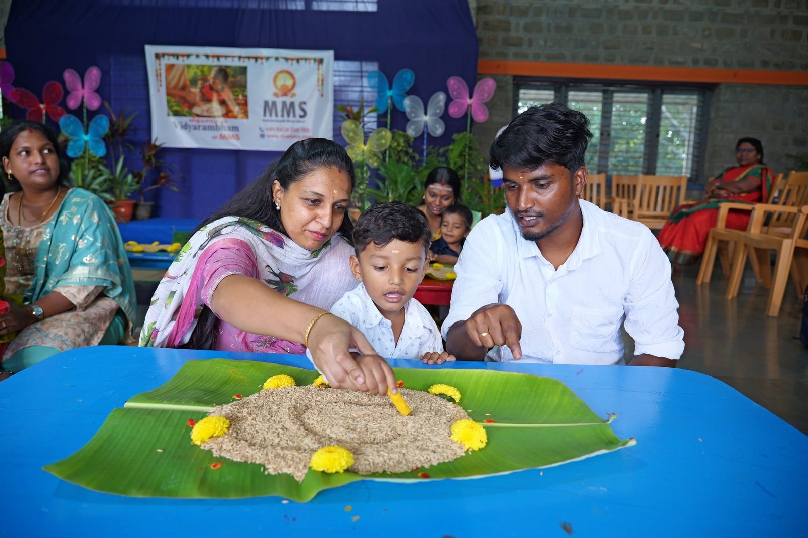 A boy writes his first letters in rice on a banana leaf, guided by his parents, at MMS Vidhyarambam