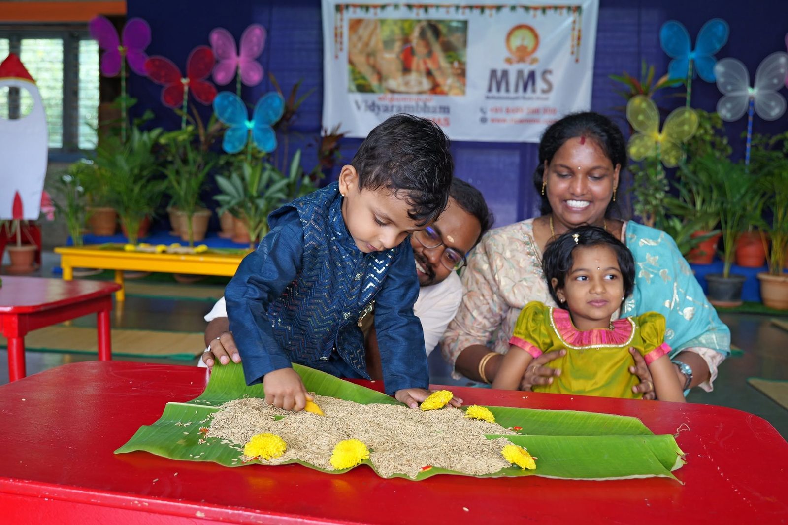 A boy in a blue kurta traces letters in rice on a banana leaf while a girl and her mother look on