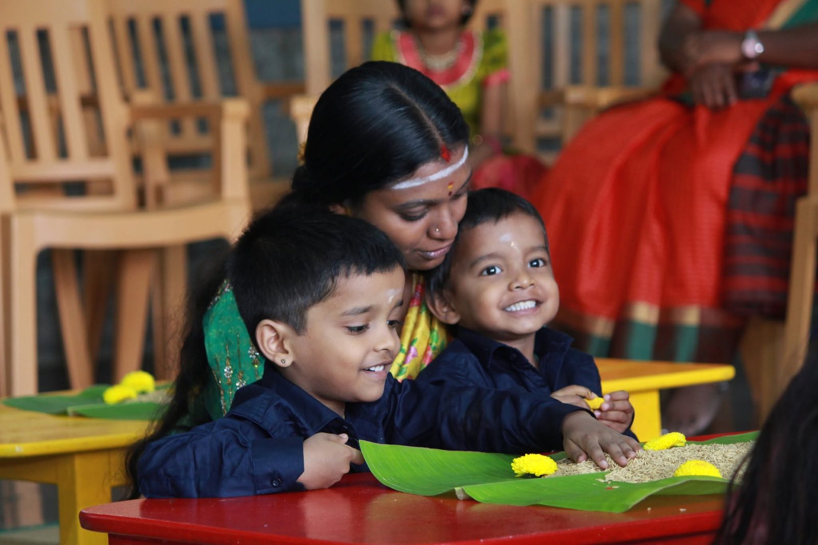 Two young boys smile as they write in the rice tray with an elder beside them